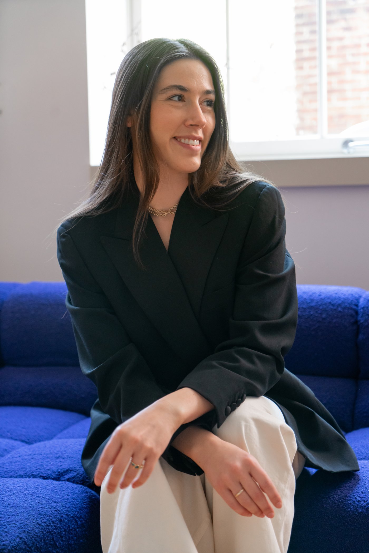 Caroline Dilsheimer sitting on a blue sofa, smiling, wearing a black blazer and cream trousers