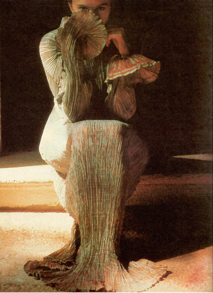 Woman in a pleated metallic Fortuny-style gown seated in dramatic light